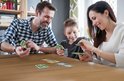 Een gezin speelt Skip-Bo Junior aan een houten tafel. De vader met een blauw geruit shirt, de moeder met een witte trui en hun zoon spelen kaarten.