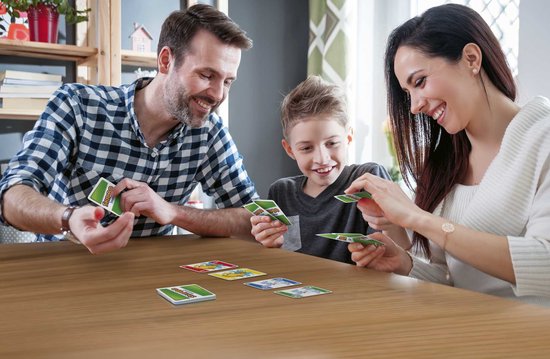 Een gezin speelt Skip-Bo Junior aan een houten tafel. De vader met een blauw geruit shirt, de moeder met een witte trui en hun zoon spelen kaarten.