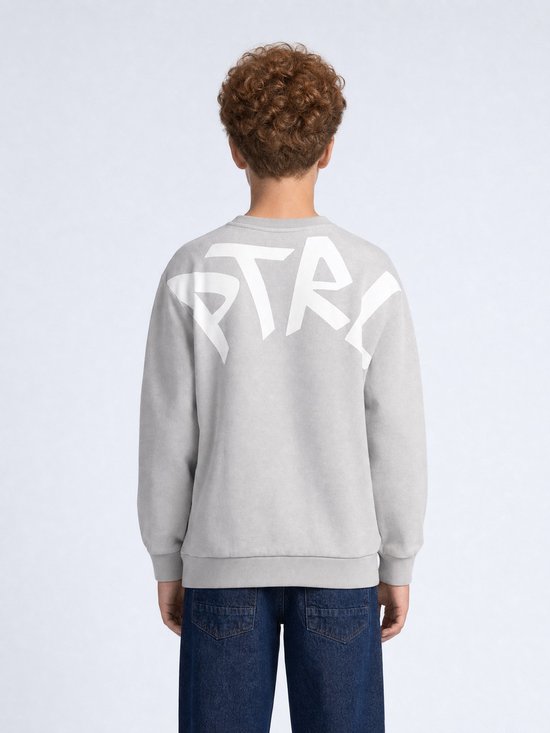 Achterkant jongen met grijze sweater met grote witte 'PTRL' print op de rug, blauwe jeans.