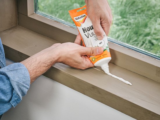 Handen gebruiken een Alabastine Houtvuller tube om witte vulmiddel op een houten vensterbank aan te brengen.