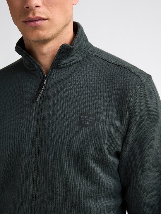 Close-up van een man in donkergroen vest met geborduurd logo op de borst.