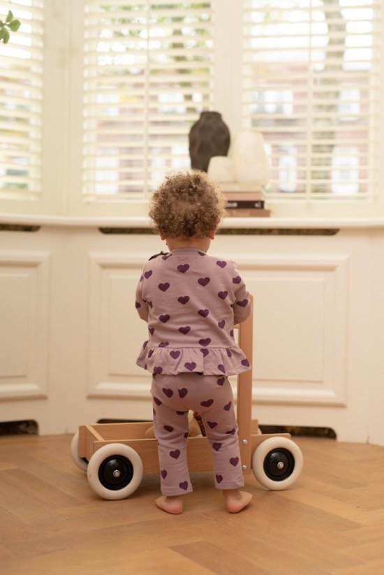 Peuter van achteren gezien in roze set met paarse hartjes, staand bij een houten loopwagen.