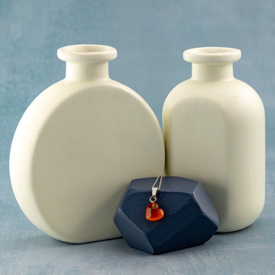 Zilveren ketting met hartvormige, oranje carneool hanger, staand op een donkerblauwe steen tussen twee crèmekleurige vazen.