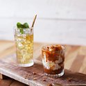 Deux verres sur une planche en bois : un verre de boisson jaune et glace, un autre avec un dessert à la sauce caramel.