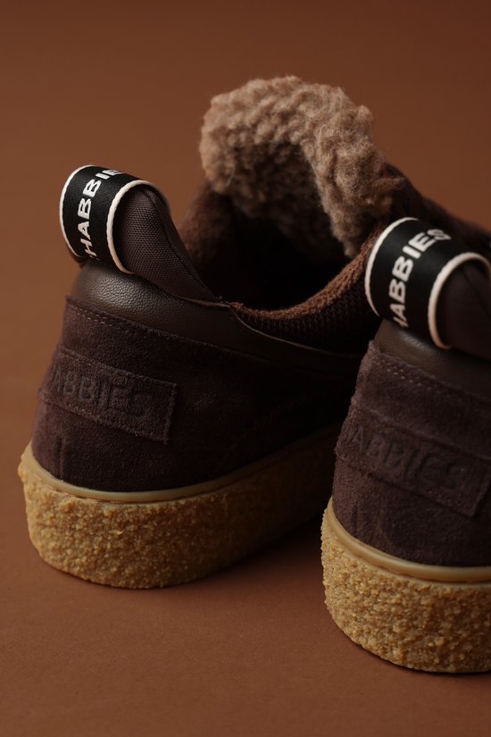 Detail van de achterkant van donkerbruine suède sneakers. De schoenen hebben een crèmekleurige zool, teddy voering en zwarte "SHABBBIES" logo bandjes op de hiel.