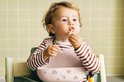 Een baby met rood-wit gestreept shirt en roze slab eet spaghetti in een kinderstoel.
