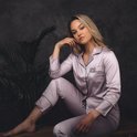Model met blond haar, zittend, draagt een lichtroze satijnen pyjamaset met zwarte biezen. Ze leunt op haar hand.