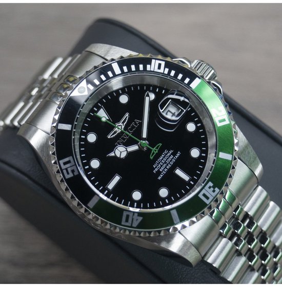 Automatisch herenhorloge met zwarte wijzerplaat, groene lunette en zilverkleurige band op standaard.