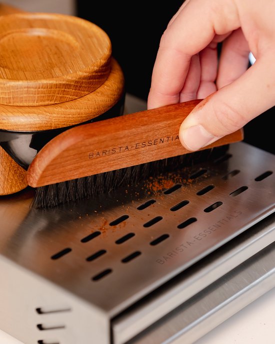 Een hand met een houten borstel met het opschrift 'BARISTA-ESSENTIALS' veegt koffiedik van een metalen rooster.