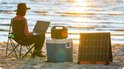Vrouw op strand met laptop, koelbox, Jackery powerstation en zonnepaneel bij zonsondergang.