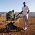Een man in een lichte outfit duwt een olijfgroene kinderwagen met wieg in een dorre, rotsachtige omgeving.