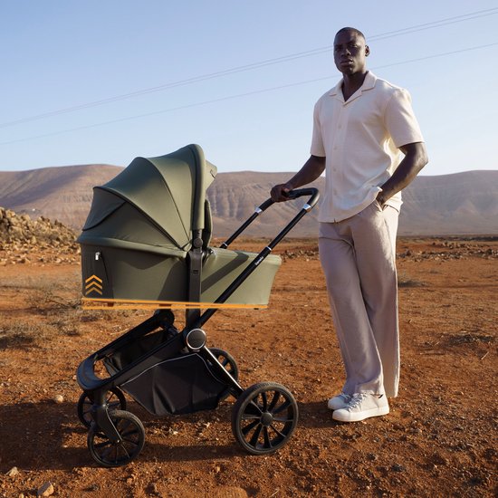 Een man in een lichte outfit duwt een olijfgroene kinderwagen met wieg in een dorre, rotsachtige omgeving.