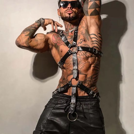 Man met zwart kunstleren harnas, tattoos, zonnebril, poses met armen.