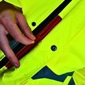Buitenkant van een felgele jas met een rode rits en grijze reflecterende details. Een hand opent de rits.