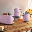 Roze SMEG broodrooster, waterkoker en citruspers op een houten tafel met sinaasappels.