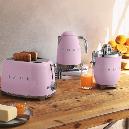 Roze SMEG broodrooster, waterkoker en citruspers op een houten tafel met sinaasappels.