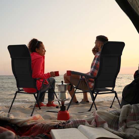 Man en vrouw zitten in zwarte campingstoelen op strand bij zonsondergang, met campinggasstel.