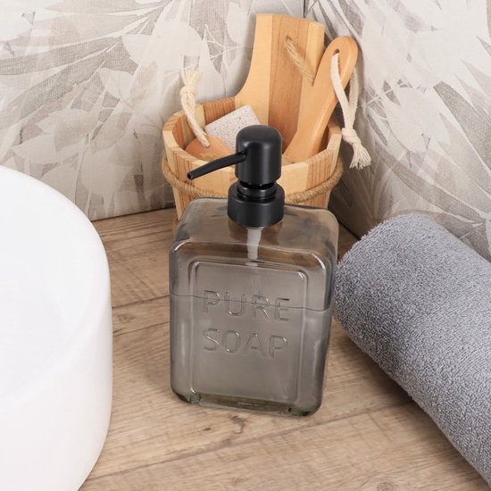 Grijze glazen zeeppomp met zwarte pomp en tekst "PURE SOAP". Naast een witte wasbak, handdoek en houten badaccessoires.