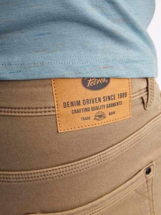 Close-up van het leren Petrol Industries 'Denim Driven Since 1989' label op een bruine chino.