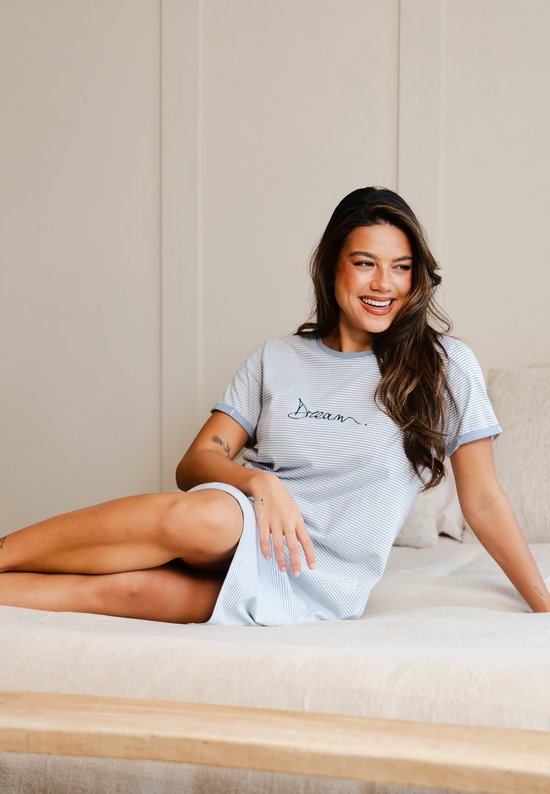 Vrouw in lichtblauw gestreept nachthemd met korte mouwen en de tekst "Dream", zittend op een bed.