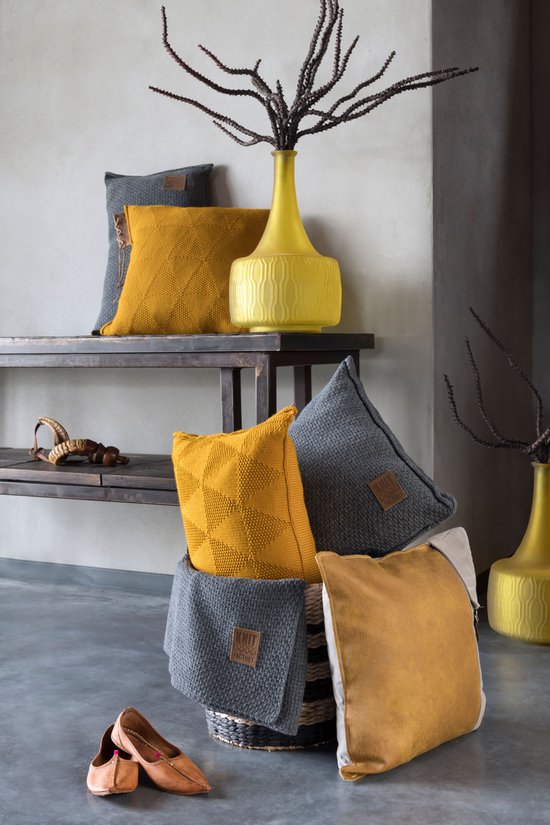Coussins gris et moutarde et un plaid avec différents motifs tricotés, à côté d'un vase jaune et de chaussures marron.
