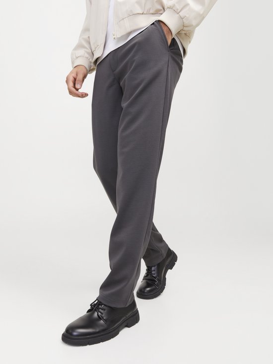 Close-up van de benen van een man, die een donkergrijze chino draagt met een beige jasje en zwarte schoenen.