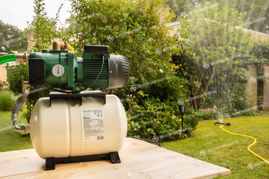 Groene waterpomp met beige druktank op een houten tafel in een tuin. Met sproeiende tuinslang op de achtergrond.