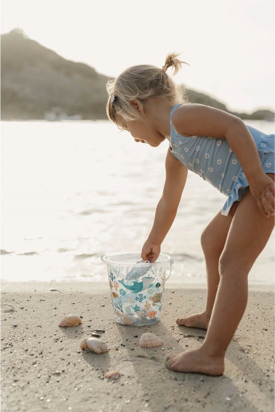 Meisje op het strand dat in een lichtblauwe emmer met zeelevenprint kijkt.