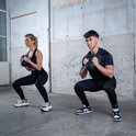 Man en vrouw doen squats met zwarte kettlebells in een industriële gymzaal.