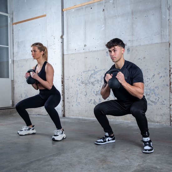 Man en vrouw doen squats met zwarte kettlebells in een industriële gymzaal.