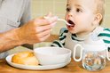 Baby met slabbetje en drinkbeker krijgt eten van witte lepel.
