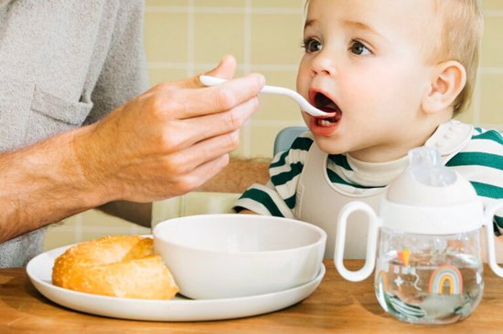 Baby met slabbetje en drinkbeker krijgt eten van witte lepel.