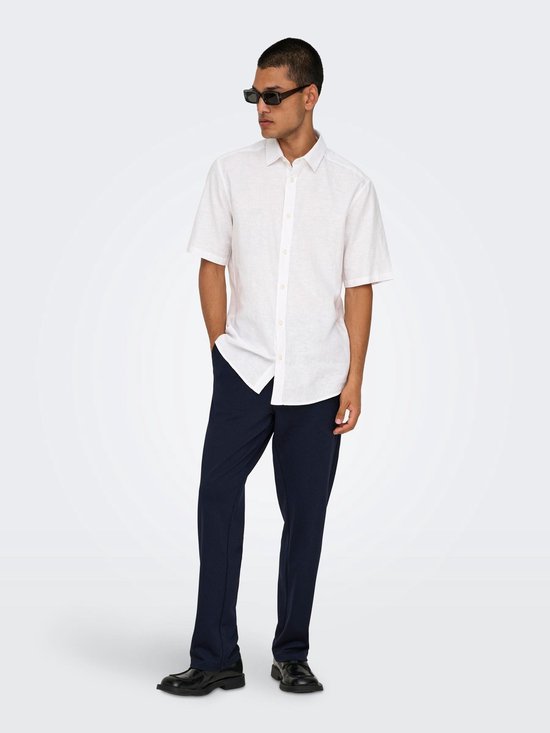 Man met donkere broek en wit hemd, handen in zakken, met zonnebril.