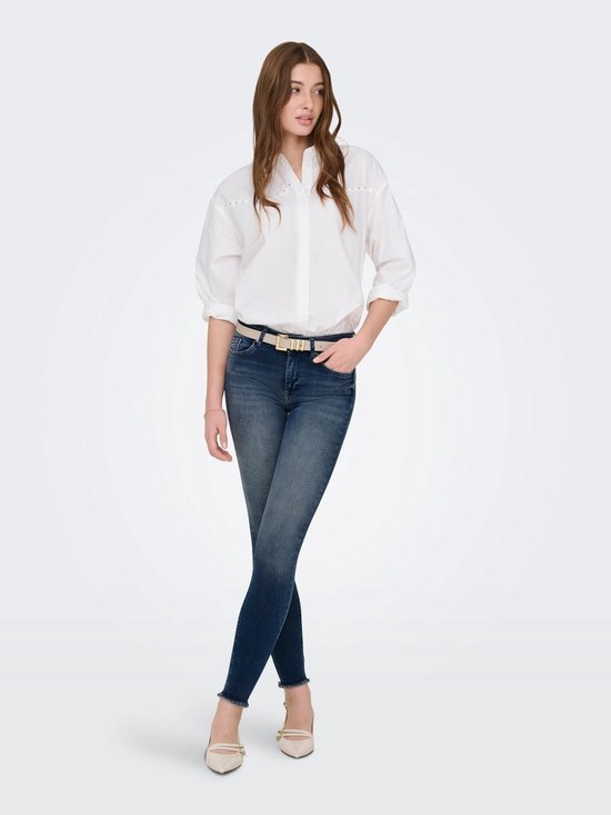 Model in witte blouse, beige riem en donkerblauwe skinny jeans met rafelige zoom.