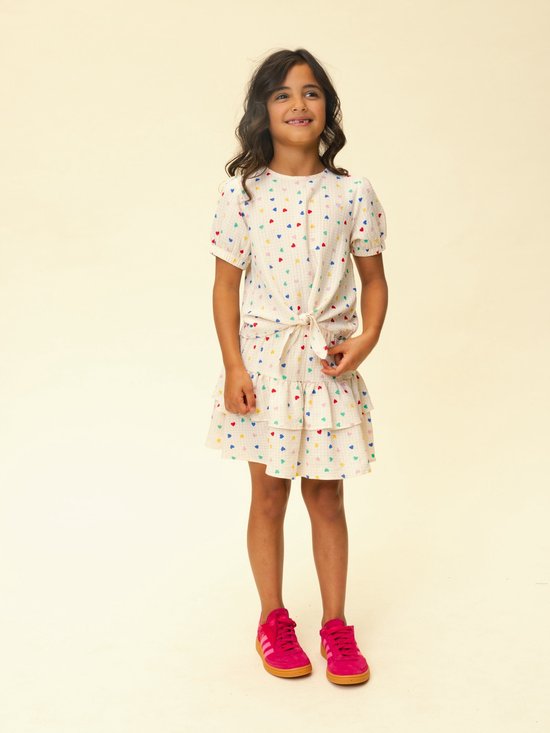 Meisje met lange bruine haren, in een top met hartjesprint en mintgroene ruchesrok, met roze sneakers.