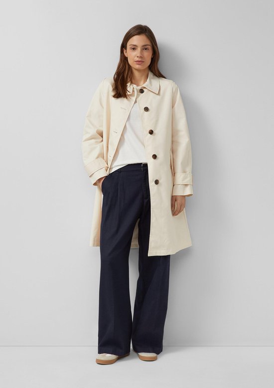 Vrouw in crèmekleurige s.Oliver trenchcoat, wit T-shirt en navy broek, staand.