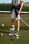 Man bij een tennisnet met witte broek, gestreepte polo, marineblauwe trui om de nek en sokken met ruitpatroon.
