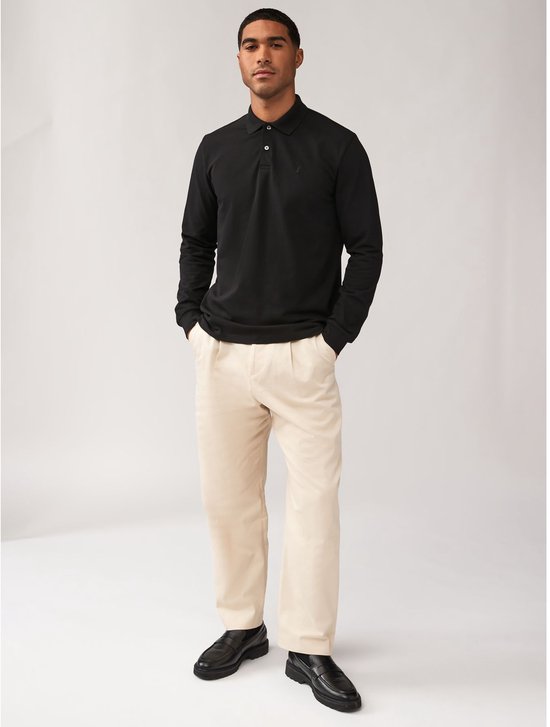 Man met handen in de zakken, draagt een zwart poloshirt met lange mouwen en beige broek.