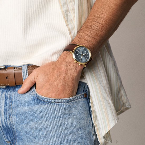 Gouden Fossil horloge met blauwe wijzerplaat en bruine leren band gedragen bij een spijkerbroek.