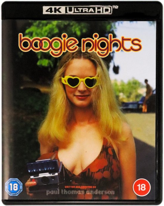 Een vrouw met blonde haren en een oranje jurk met hartvormige gele zonnebril, houdt een camera vast.