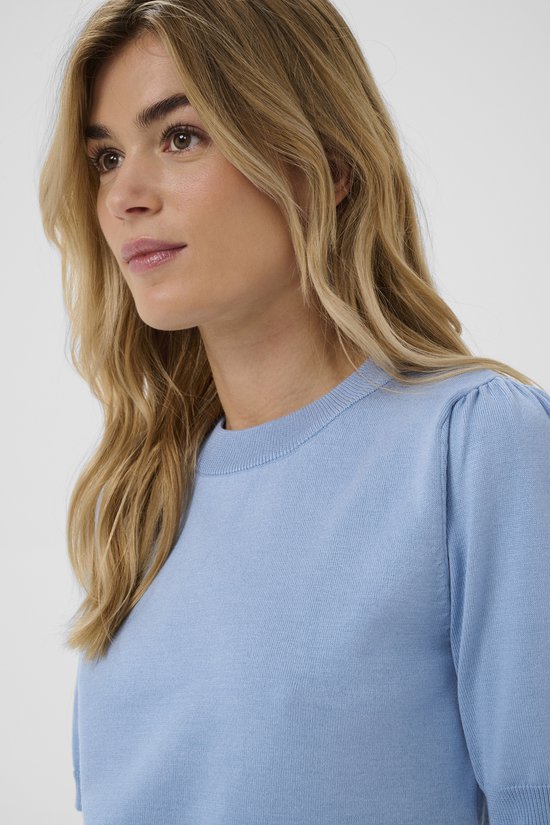 Een blonde vrouw met een lichtblauwe trui met korte, gepofte mouwen en een ronde hals.