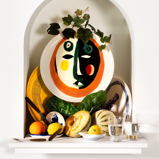 Een grote serveerschaal met een gezicht met zwart-witte helften, een oranje rand en groene ogen, in een nis met fruit.