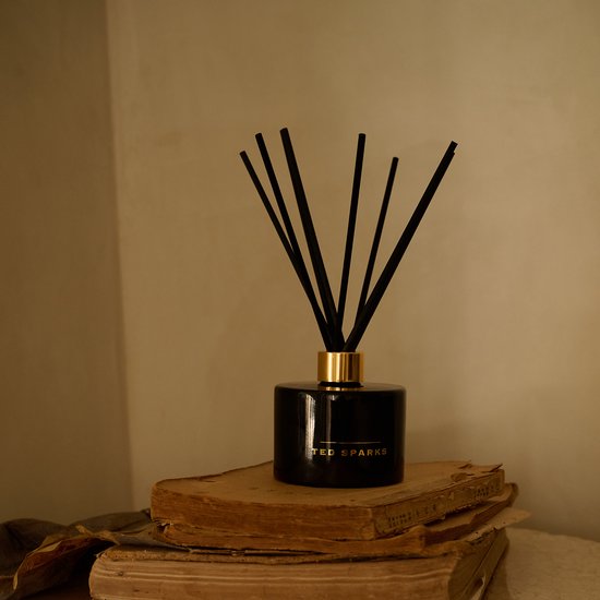 Zwarte glazen diffuser van Ted Sparks met gouden dop en zwarte stokjes, op gestapelde oude boeken.
