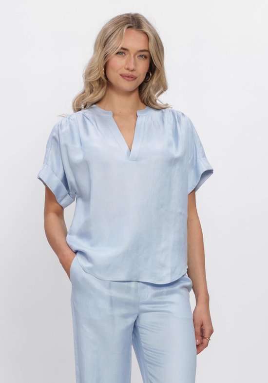 Model in lichtblauwe blouse met korte mouwen en V-hals. De blouse heeft plooien bij de schouders en is gecombineerd met een bijpassende broek.