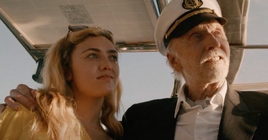 Een blonde vrouw in een gele jurk, met een zonnebril op haar hoofd, leunt op de schouder van een oudere man in een kapiteinspet.