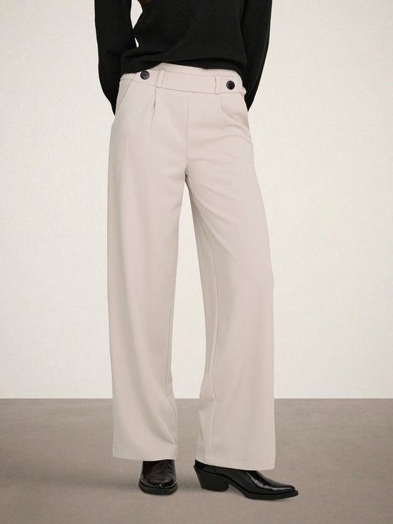 Lichtbeige wijde broek met plooitjes en zwarte knopen aan de voorkant, gedragen door een model.