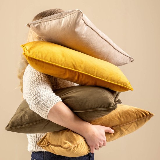 Een vrouw met blond haar, gekleed in een gebreide trui, die een stapel fluwelen kussens draagt in verschillende tinten groen, geel en beige.