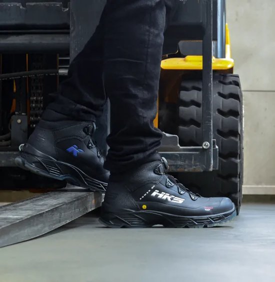 Voeten in zwarte HKS werkschoenen, een met blauw K-logo, staand op een steiger.