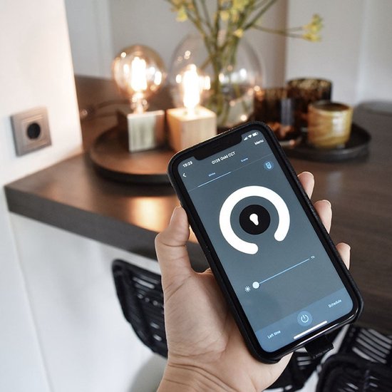 Hand houdt smartphone met lichtinstellingen, met gedimde lampen op achtergrond.