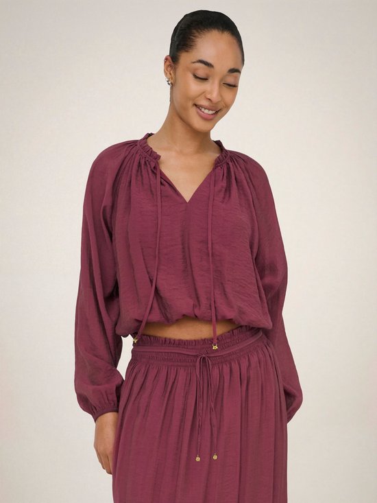 Model draagt een bordeauxrode crop top met pofmouwen en een v-hals met strikdetail.